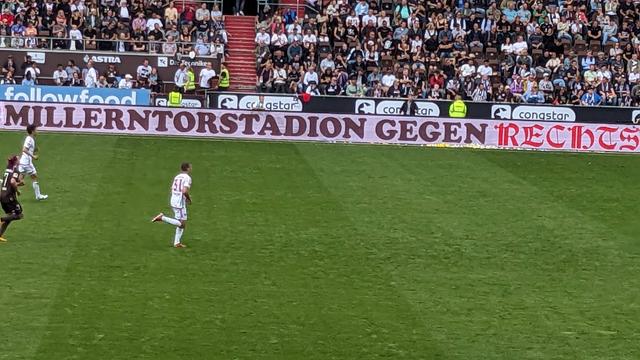 Drei Fußballer auf dem Rasen. Auf der Bande ist der Schriftzug "MILLERNTORSTADION GEGEN RECHTS" zu lesen. Im Hintergrund die Zuschauenden auf der Haupttribühne. 