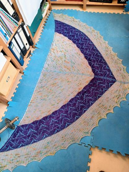 A large knit triangle shawl blocking on a blue blocking mats.