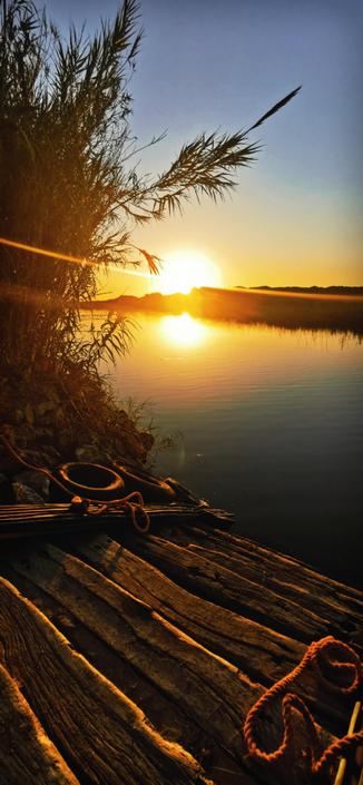Eine alte Holztreppe vor einem See mit Schilf. Im Hintergrund geht die Sonne unter. 