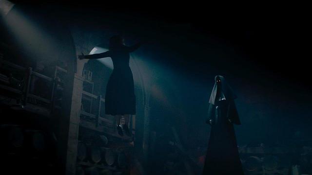 A weird creepy image from THE NUN II, showing a womanly figure floating in front of a stream of light from a window, with a demon nun watching her from beneath