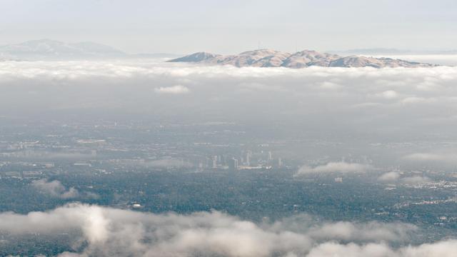 Downtown San Jose, California, seen through a clear strip of sky from many miles away up high. A layer of fog separates it from Mission Peak in the background and Mount Diablo in the distance on the left. Much human suburban development otherwise. 