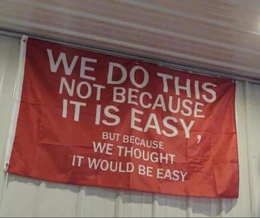 Red flag with white lettering hung up high on a wall that says 
We to this not because it is easy,
but because we thought it would be easy"