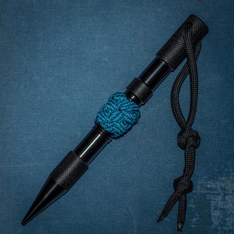 A photograph of a black metal marlinspike/case with a sailor's knife lanyard knot and double wall knot on the paracord lanyard and a 4-bight, 5-lead Turk's Head tied around the spike in bicolor blue cord. 
