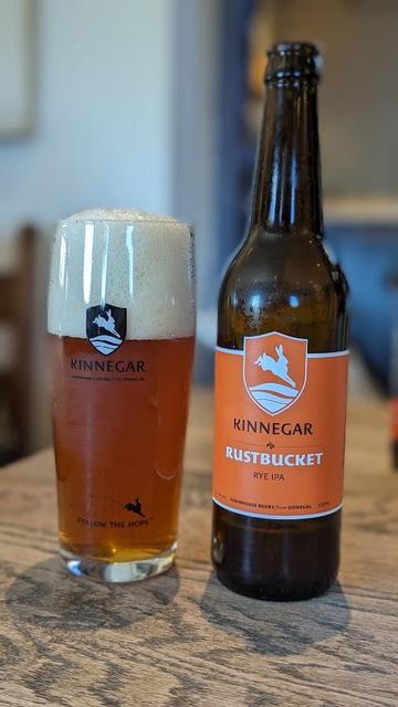 Auf einer hölzernen Tischoberfläche steht recehts eine Flasche Bier und links ein volles Glas Bier mit einer Schaumkrone. Auf der Falsche ist ein oranges Etikett mit der Aufschrift "Kinnegar Rustbucket Rye IPA".