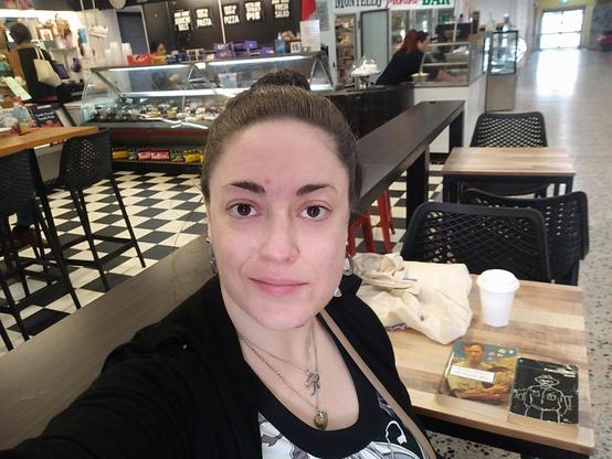 Colour; landscape orientation: A 30-something Caucasian woman (me) with brown hair in a bun and wearing black, white, grey and brown smiles a little bit, braving public view despite her blemished complexion while sitting at an Italian delicatessen with her coffee, Patrick White novels, and calico shopping bag that contains trinkets and a Star Trek paperback.