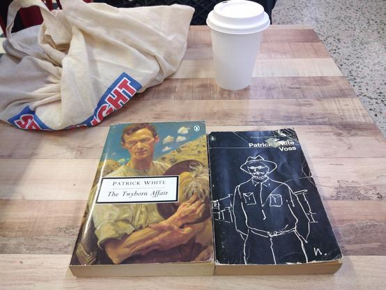 Colour; landscape orientation: Patrick White's "The Twyborn Affair" paperback on the left and his "Voss" on the right, with both a crumpled calico carry bag and a biodegradable takeaway coffee cup in the background.