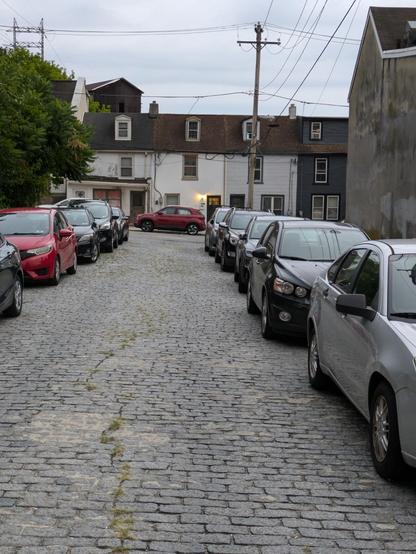Photo: Standing in the middle of a narrow cobblestone street lined on both sides by cars facing the photographer. At the far end of the street is a row of connected tiny houses, each with a single window on the second floor and a single dormer window protruding from the roof above the second floor.