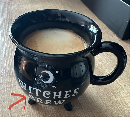 Tasse in Form eines Hexenkessels mit Kaffee darin. Roter Pfeil zeigt auf die Aufschrift „Witches brew“