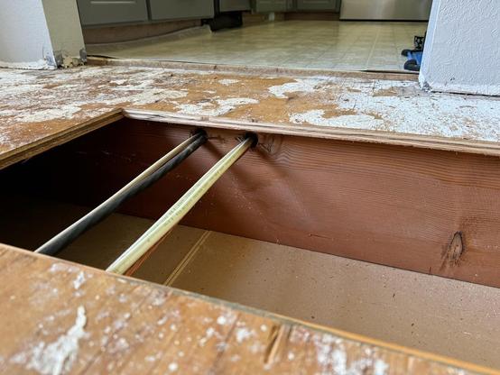 An open floor joist bay during a home remodel. Electrical wires run perpendicular to the joist through holes that are extremely close to where the subfloor plywood gets nailed on top.