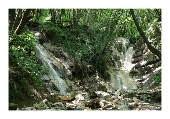 The image shows a serene scene of a natural waterfall cascading over rocky terrain, surrounded by lush green foliage in a forested area