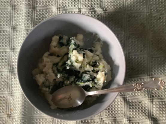 Round grey bowl with mashed potatoes with green bits of kale and a spoon sticking out over a light green tablecloth.