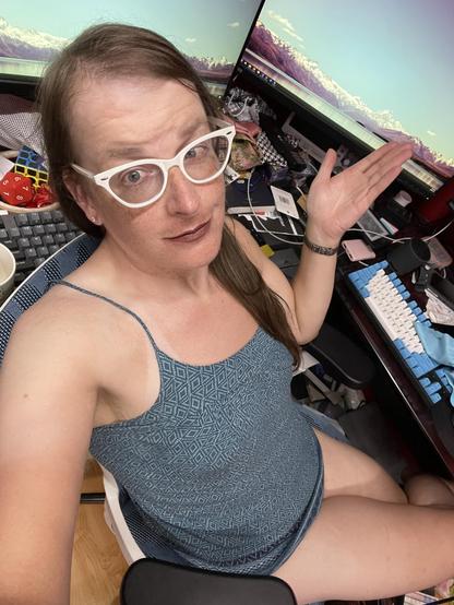 Cailín is sitting at her desk. She’s wearing white cats eye glasses, mauve lipstick, and a teal strappy tank top with a geometric pattern on it 
