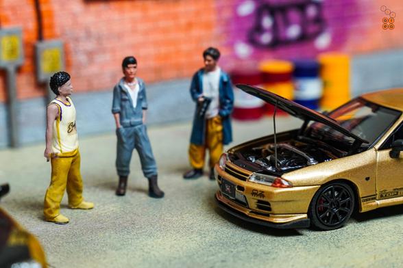 3 guys stand beside Toyota Supra A80 by Time Micro and Nissan Skyline GT-R VR32 by Facal Horizon, both with Top Secret Gold livery in 1:64 scale on AREA 164