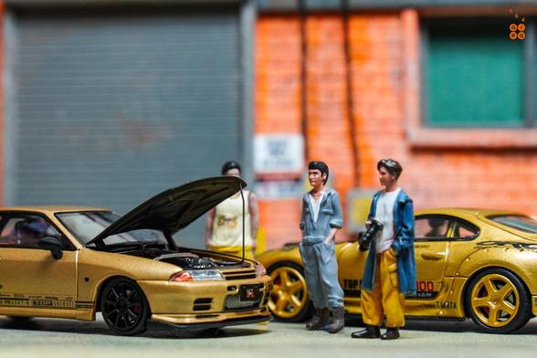 3 guys stand beside Toyota Supra A80 by Time Micro and Nissan Skyline GT-R VR32 by Facal Horizon, both with Top Secret Gold livery in 1:64 scale on AREA 164