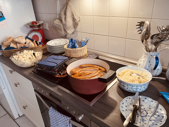 Auf einer Küchenzeile ist ein kleines Buffet mit Nudel, Krautsalat, Würstchen und Brötchen aufgebaut.