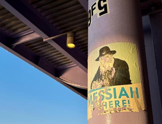 A faded and worn down flyer on a purple steel column under an elevated highway. The flyer depicts a old rabbi with the messiah is here underneath 