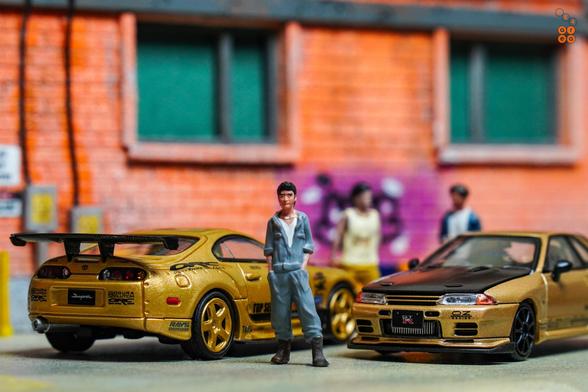 A guy stand in front of Toyota Supra A80 by Time Micro and Nissan Skyline GT-R VR32 by Facal Horizon, both with Top Secret Gold livery in 1:64 scale with 1:64 figure on AREA 164