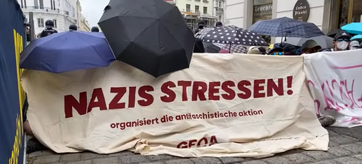 Zu sehen ist ein Transparent mit der Aufschrift "NAZIS STRESSEN!"
Darüber einige Regenschirme.