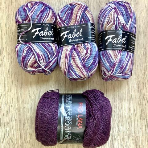 3 balls of variegated yarn in lilacs and purples, and one eggplant colored ball of yarn on a light wooden background.