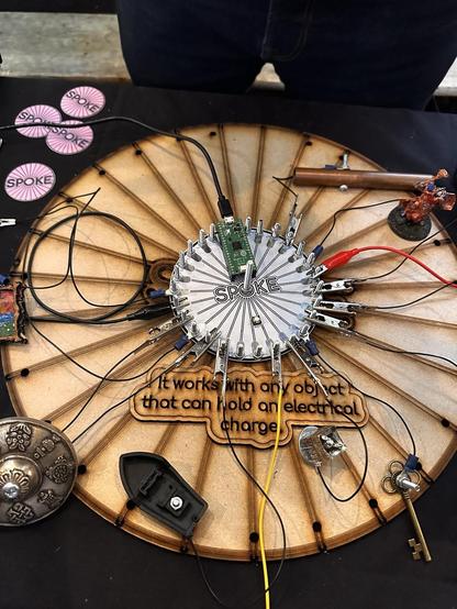 Spoke 26 capacative touch sensor MIDI controller with cables attached to small objects resting on a wooden board. Text says it works with any object that can hold an electrical charge.