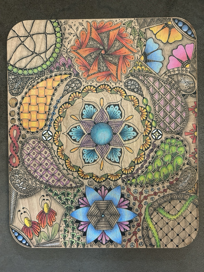 Wooden board with zentangle-style drawings in black pen and coloured with pencil. Starting with a blue and purple mandala in the inside, with tear drops surrounding with grid work tangles inside, circles on the top left and bottom right with sectional tangles inside, hexagon shapes on top right and bottom left with flowers inside, and swirls top and bottom middles. In between the main shapes are different tangle shapes and designs 