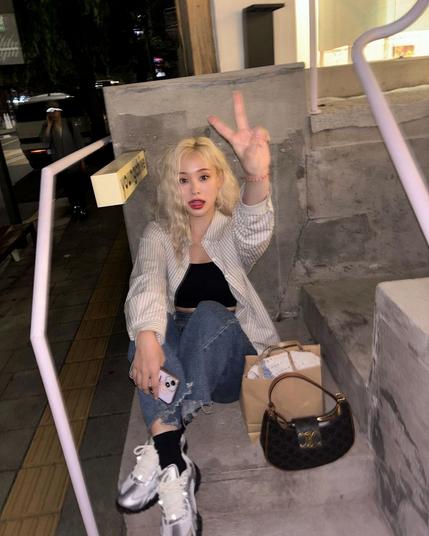 photo of gahyun (dreamcatcher) sitting on the stairs in front of a building at nighttime. she has long blonde hair, is wearing a white button down shirt, a black crop top and blue jeans. showing ✌️ to the camera.