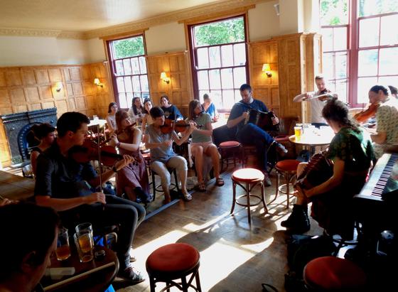 Photo of the front room of the Lamb and Flag pub in Oxford with late afternoon sunlight streaming in through the large windows. Participants of a folk session playing fiddles, flutes and melodeons are sitting in a circle and concentrating on their playing. 