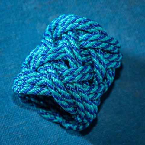 A photograph of a gaucho knot, tied in bicolor blue cord, sitting on a blue cloth surface.

