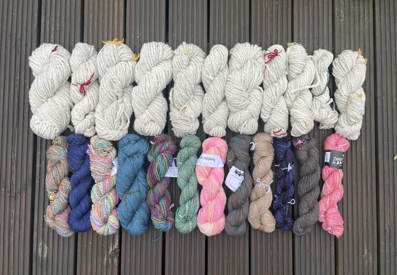 2 rows of skeins of yarn laid on a wooden deck. The top row is off white chunky, the bottom row is multicoloured and 4ply/dk
