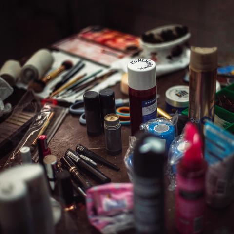 Auf einem Tisch im Theater sind verschiedene Schmink- und Theaterutensilien zu sehen: Makeup, Grundierung, Haarspray und -gel, Bandagen, Pinsel, Eyeliner und in der Mitte eine größere Flasche Kunstblut. 