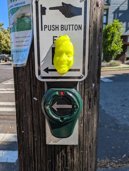 A smiling, yellow, probably male human face created through 3D-printing (I think) has been glued on to a sign that tells you to push a button below to activate a pedestrian crosswalk light, for those of us who believe that these buttons actually work.