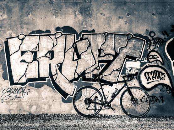 Monochrome photo of a black specialized diverge gravel bike standing next to a concrete wall with a large piece of graffiti style art on it. the graffiti art prominently features the word “ERUPT”