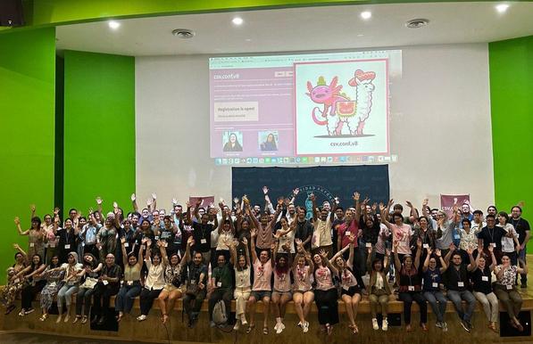Group photo of csv,conf attendees, the background is a colorful csv,conf banner.