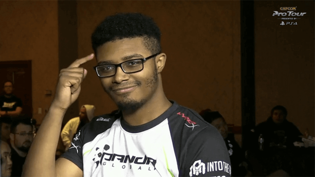 O jogador de Street Fighter Punk, dos EUA: jovem negro, de cabelo cortado baixinho dos lados, de óculos de armação quadrada, vestindo uma camiseta esportiva branca com mangas pretas. Está fazendo pose apontando para a própria cabeça, com um sorriso leve no rosto.