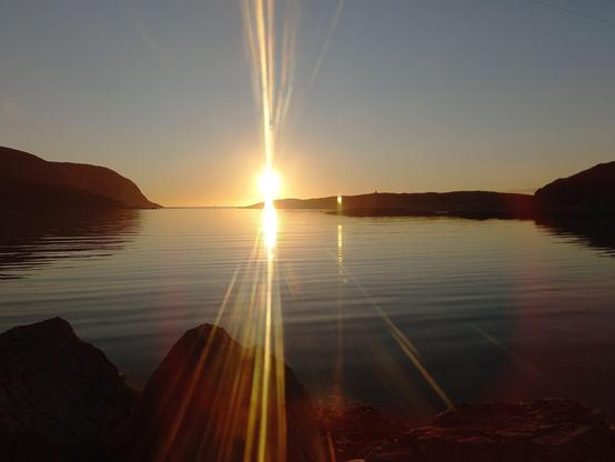 Man sieht den Fjord, das Wasser ist total ruhig, rechts und links Felsen, im diffusen Licht. Die Sonne steht knapp über dem Horizon. Ich habe das Bild durch das Camperfenster aufgenommen. Die Sonnenstrahlen werden in der Scheibe augrund von Verschmutzung und Kratzern nach oben und unten gestreut.