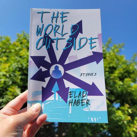 Hand holding up a book titled "The World Outside: Stories by Elad Haber." The book cover is white with blue letters and the photo background is a green bushy tree against a bright blue cloudless sky.
