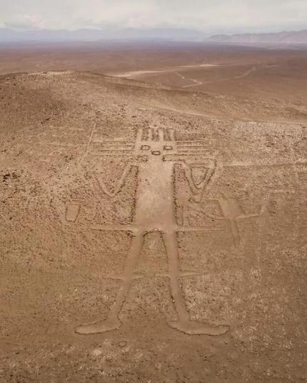 Geoglifo humanoide con brazos a ambos lados a la altura de los hombros. Cabeza cuadrada con cuatro líneas saliendo de tres de sus lados. De sus caderas y rodillas salen líneas perpendiculares.
