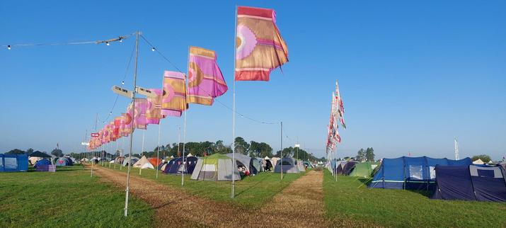 Looking up a forked path in a camp site. Colourful flags line the edges of the paths and numerous tents populate the camping areas. The sky is clear blue.