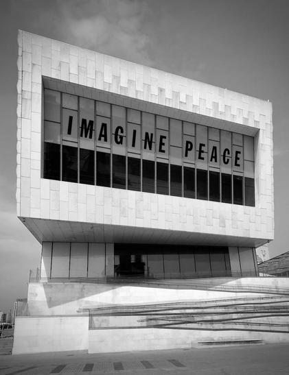 A black and white photograph showing a section of the modern building The Museum of Liverpool with the large inscription 'IMAGINE PEACE'. Each letter is placed in a separate window, which forms a huge single window occupying most of the surface area of the entire building wall. The window frame is made of sandstone blocks. The building is approached, also made of sandstone blocks, via a zigzag path with glass partitions to allow access for a wheelchair, e.g.