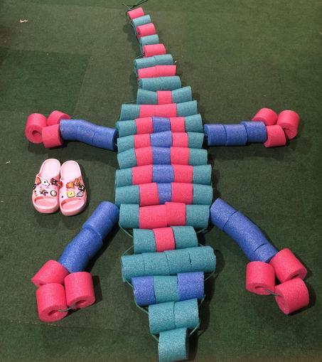 An absolute unit of a beaded lizard made of cut up pool noodles for beads. It is made of fuchsia, teal, and periwinkle noodles to give that 90s summer vibes. Next to it are size six sandals next to it to give an idea just how huge the lizard is.