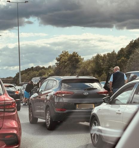 Traffic jam on a highway with people standing outside their cars. Cloudy sky and trees in the background.