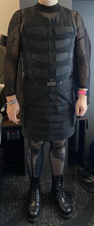 A person with a sheer longsleeve shirt, a knee length skirt and a fake bulletproof vest. They are wearing black tights and ankle high laced boots. Tattoos can be seen through the clothes on legs and arms