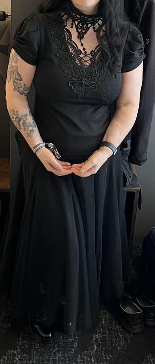 A person wearing a shirt with a V shaped neckline with laced ornaments and a ruffled, ankle long skirt. The left arm has an ivy tattoo 