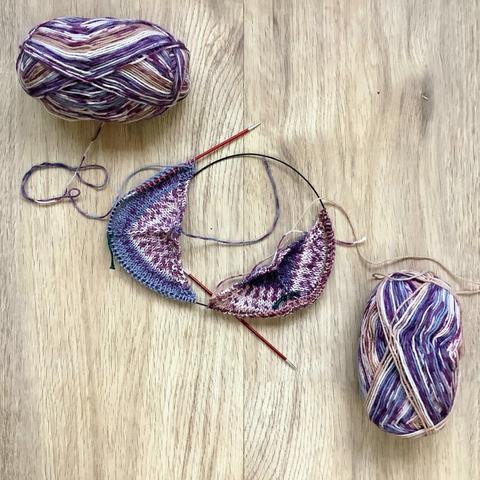 Two little knit triangles on a large circular needle. They're shown on a light wooden background, together with 2 balls of variegated yarn in various lilacs.