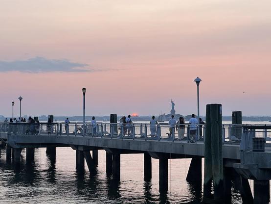 Sunset photo taken from Red Hook. The sky is hazy and pinkish and grayish. Part of Valentino Pier is in the foreground. Beyond, the sun is almost below the horizon, just a bit to the left of the Statue of Liberty.
