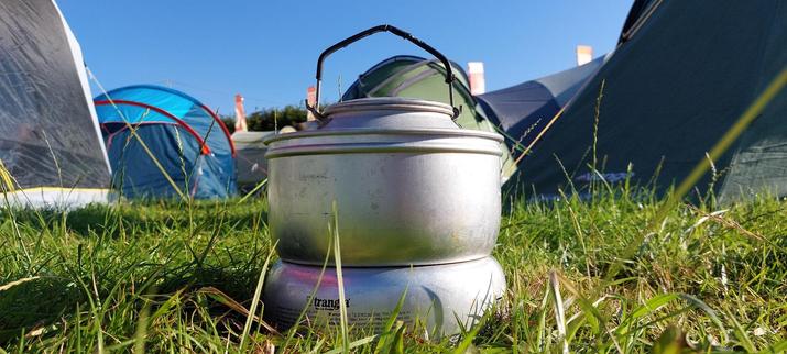 A Trangia brand camping stove and kettle on grass with tents and a blue sky in the background. 