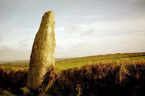 A colour photo of the menhir at Prospidnick in the parish of Sithney, Cornwall. The standing stone is on the edge of a low hedge beside a minor road and leans slightly towards the field. The green hill can be seen curving gently over the horizon towards Wendron.