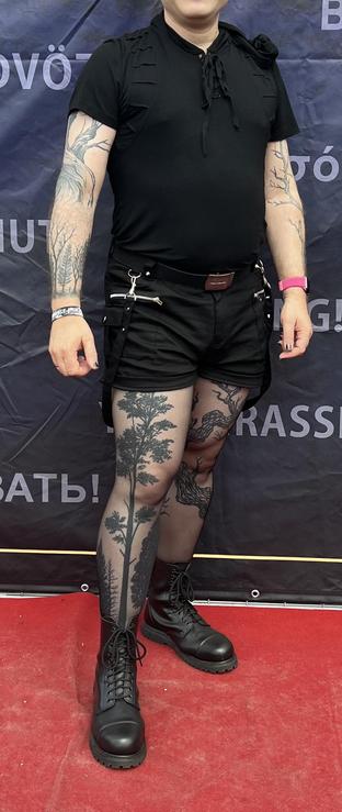 A person with a black shirt, hotpants and sheer black thighs. Arms and legs are covered with tree tattoos and they are wearing laced boots 