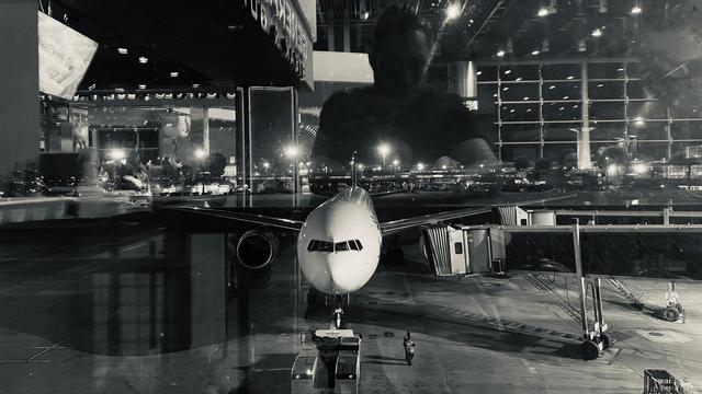 An airplane docked; through a glass; reflection shows airport interior and a silhouette, perhaps of the person who took the picture, perhaps me; black and white