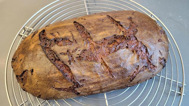 Farbfoto: Auf einem Kuchengitter liegt ein ovaler Laib Brot mit leicht glänzender Kruste, die drei Mal schräg eingeschnitten wurde, aber trotzdem auch wild eingerissen ist. 